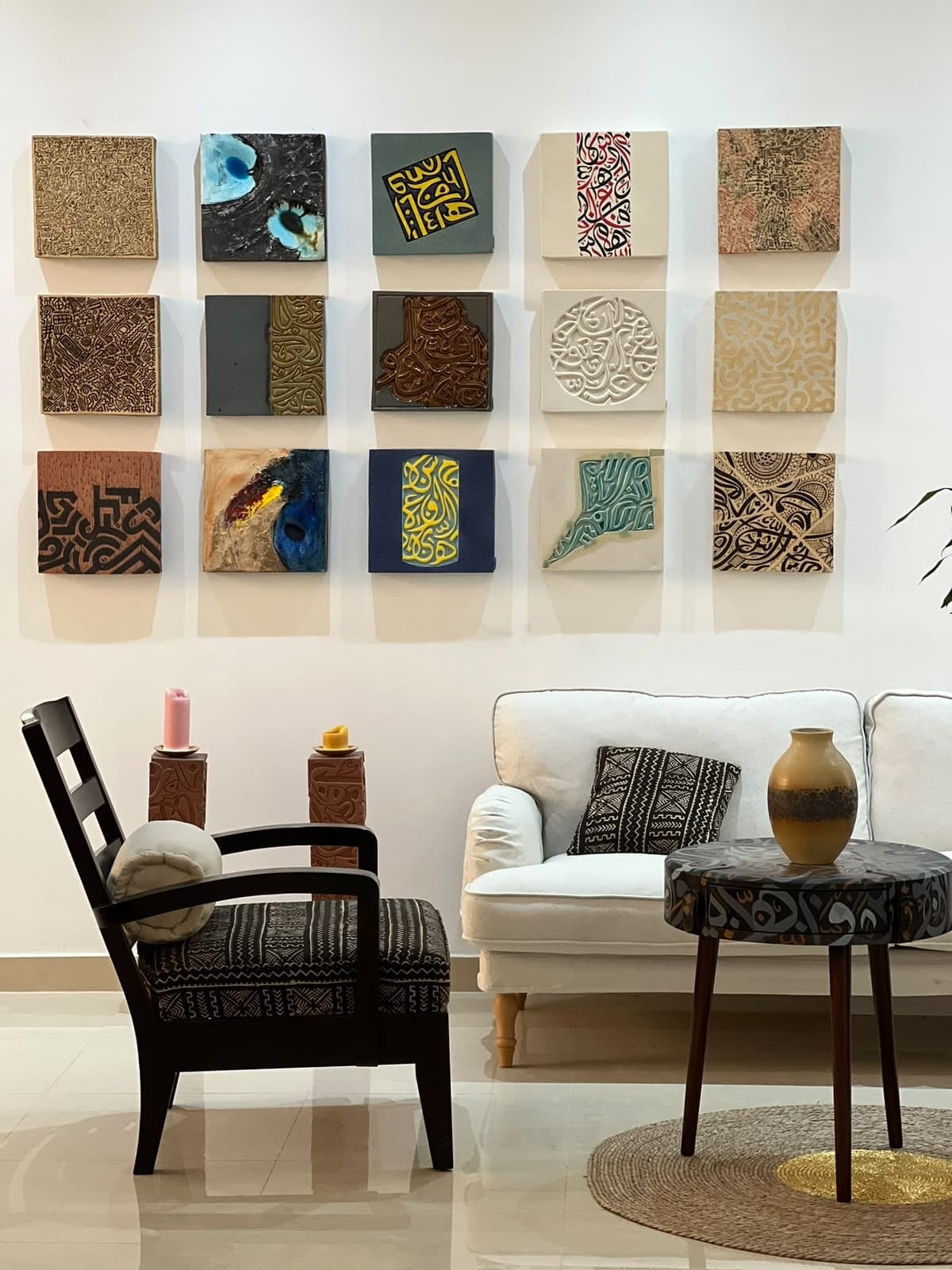 Fifteen square ceramic wall tiles with calligraphic, patterned and glazed surfaces hung in a grid above a seating area