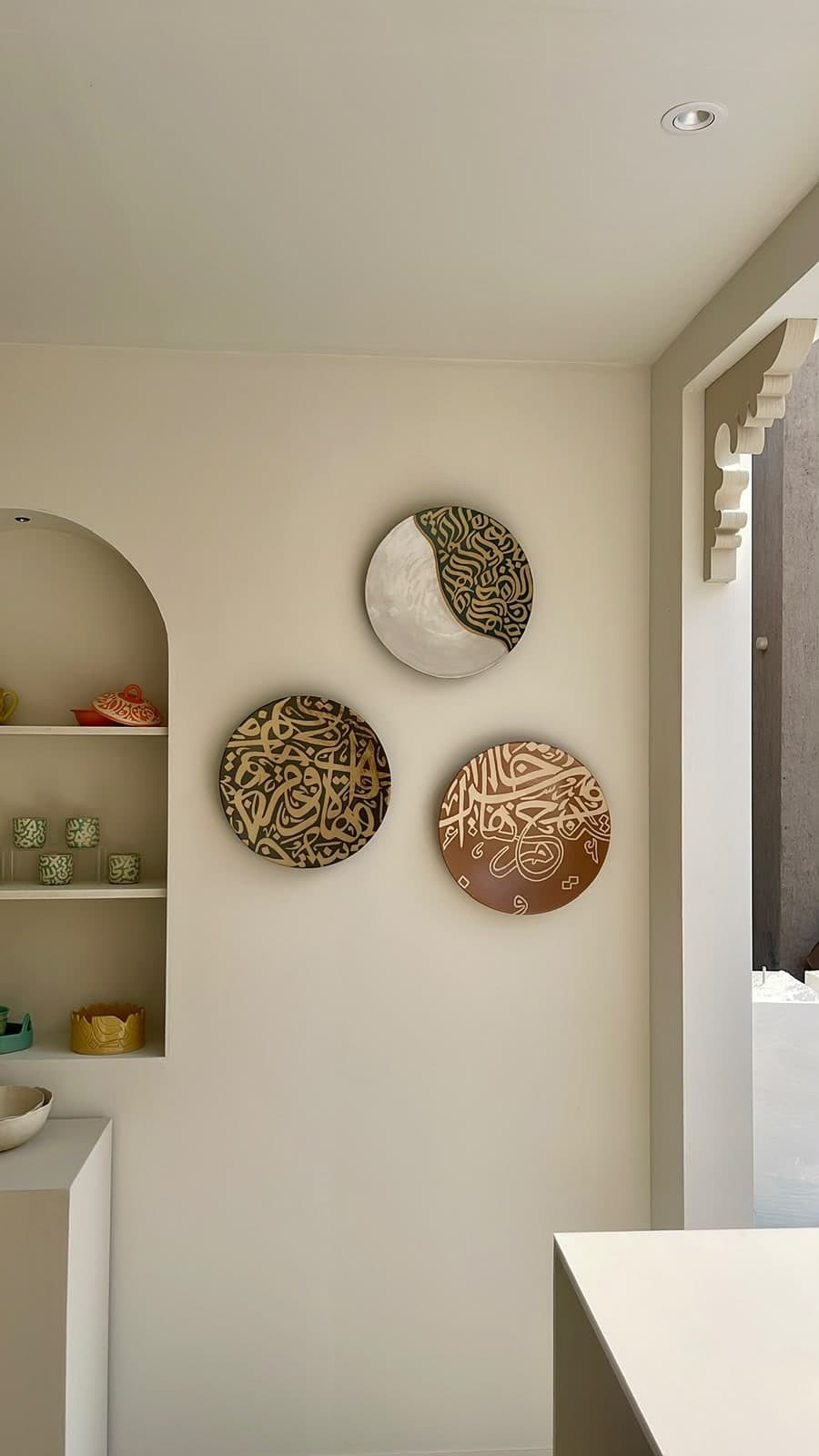 Three round calligraphy wall plates in green, black and terracotta on a cream gallery wall beside an arched shelf