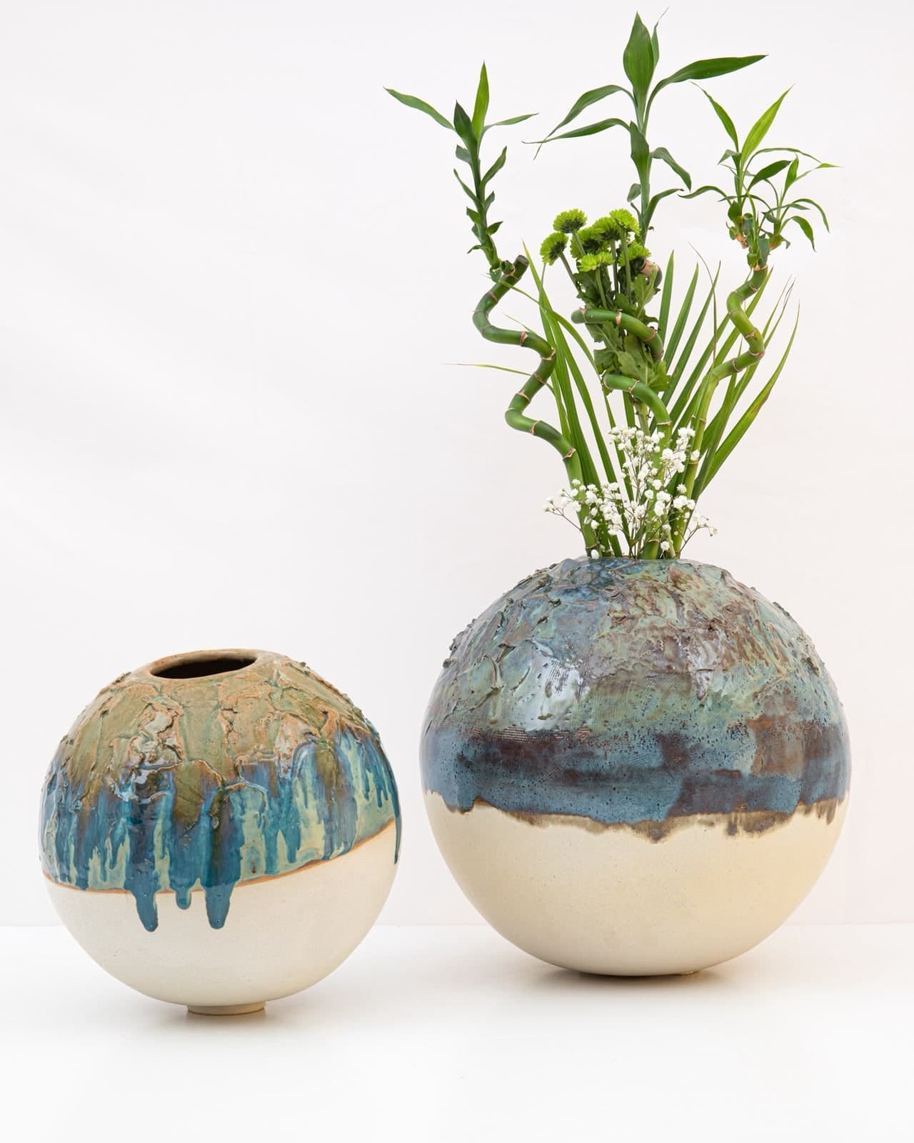 Two round ceramic moon jars with textured blue and green drip glaze over raw sand-coloured clay; the larger holds bamboo stems and white flowers