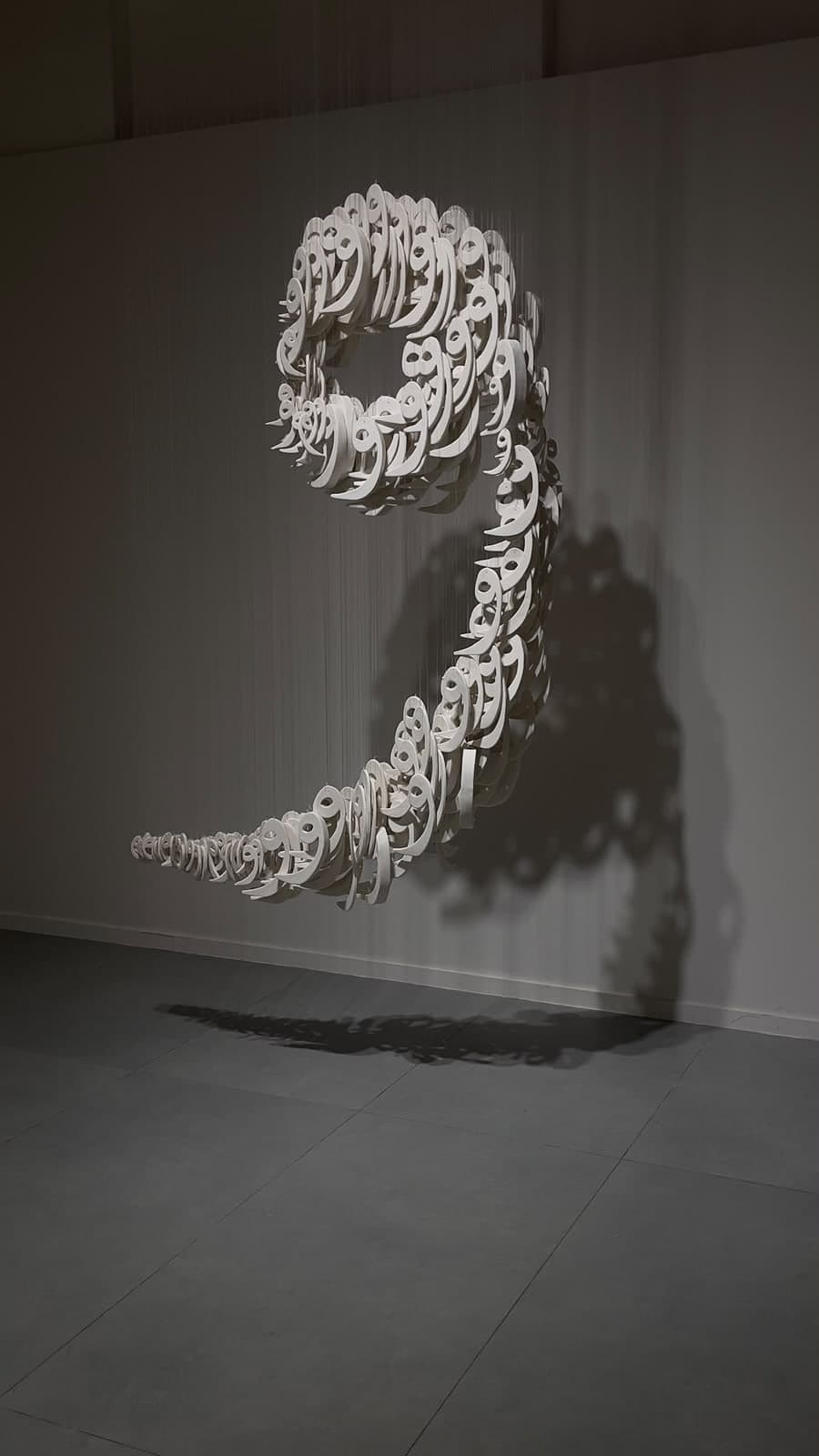 Suspended installation of hundreds of white ceramic Arabic letterforms forming a large sweeping curve, casting shadows on a gallery wall